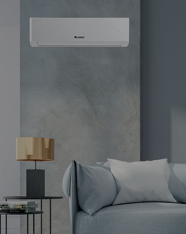 Nowoczesny salon ma zamontowaną na ścianie jednostkę klimatyzacyjną nad turkusową sofą. Na sofie leży beżowa poduszka, a na stole, obok której leżą książki, stoi złota lampa. Ściana jest pomalowana na stonowany odcień szarości, co tworzy elegancką i chłodną atmosferę.